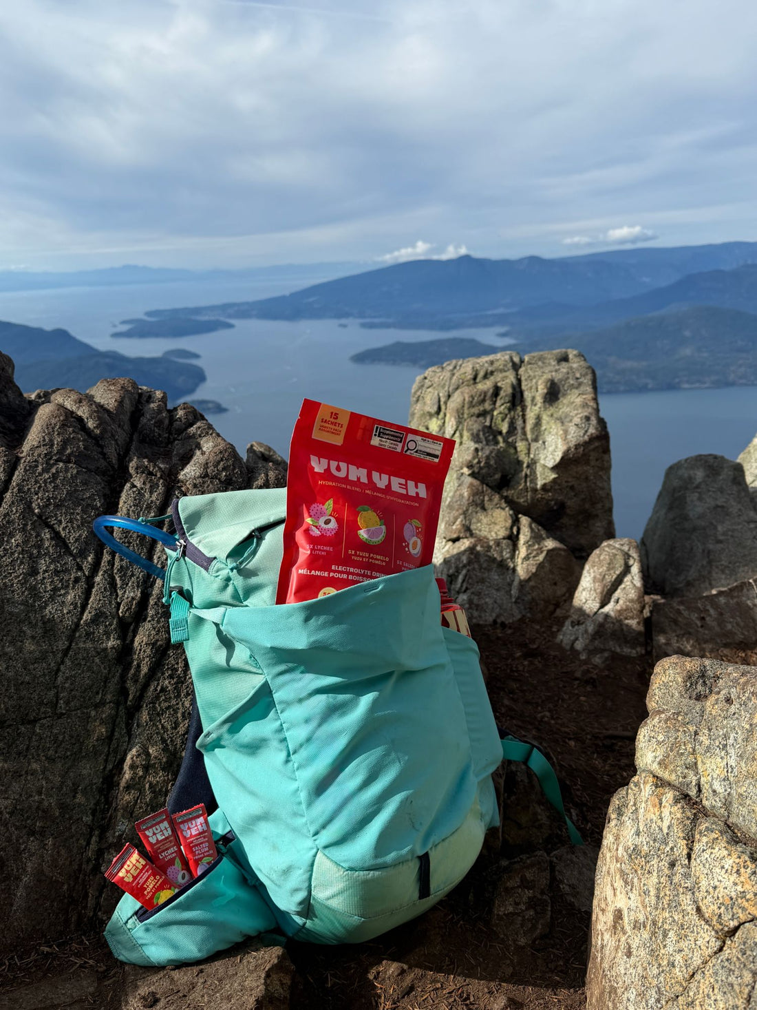 YumYeh electrolyte pouch and stick packs tucked into a teal hiking backpack on a rocky summit above the ocean