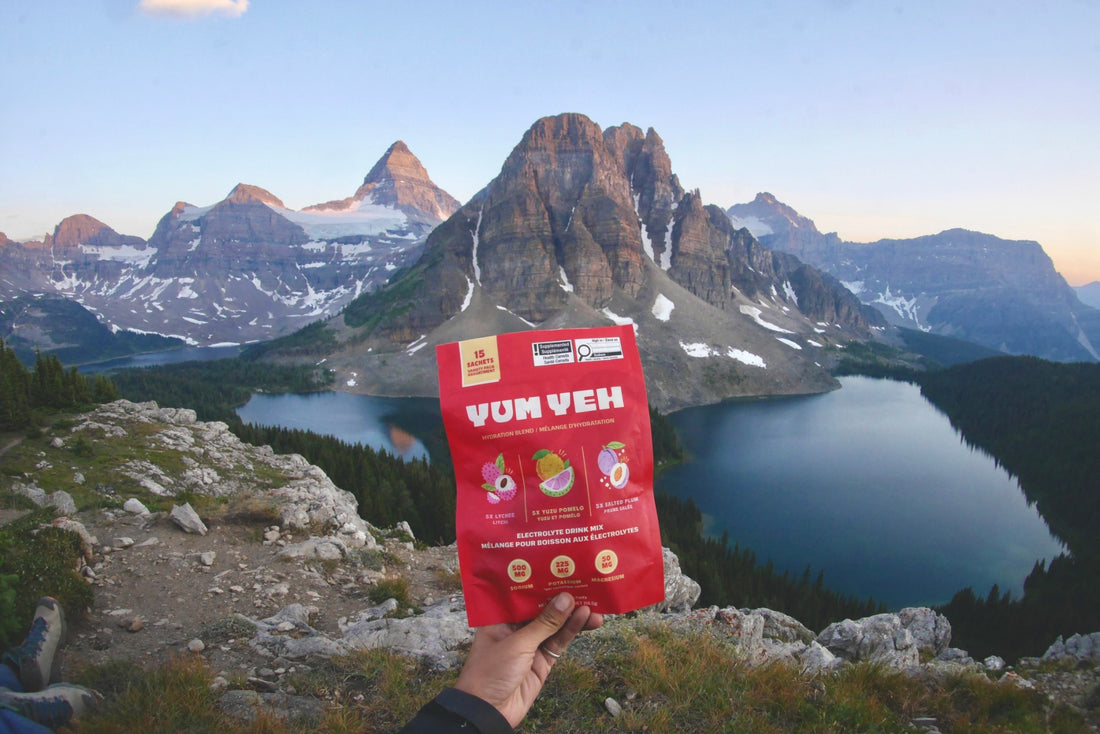 Hand holding the YumYeh electrolyte variety pack pouch in front of a mountain lake at sunrise