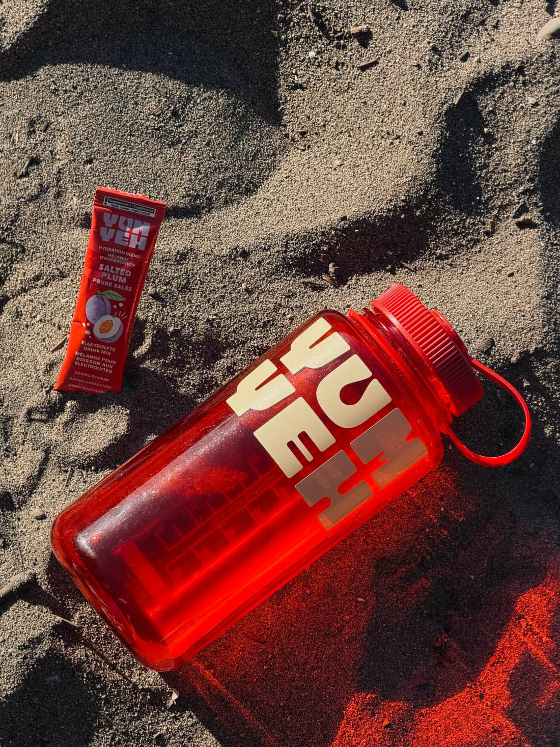 YumYeh salted plum electrolyte stick pack beside a red YumYeh water bottle on sand