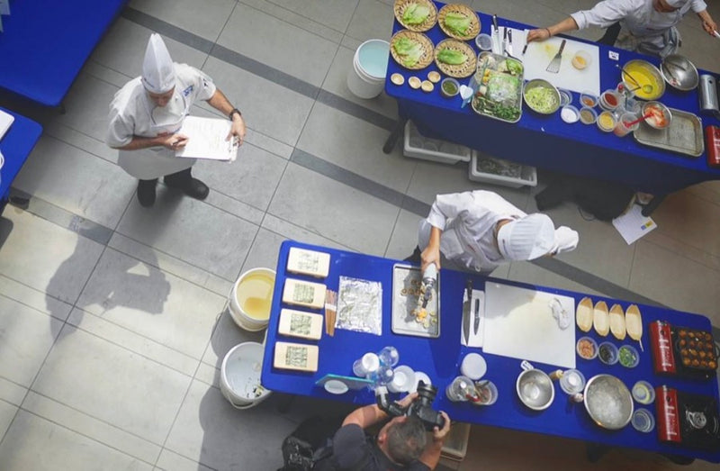 A cooking competition seen from above, with chefs at blue tables and a judge taking notes