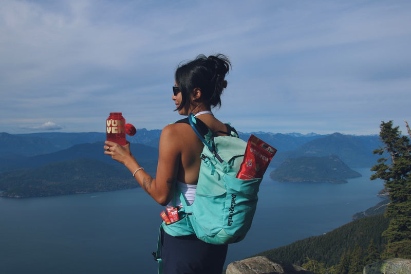 Jennifer on a summit above the ocean holding a YumYeh water bottle, with a pouch and sticks in the backpack